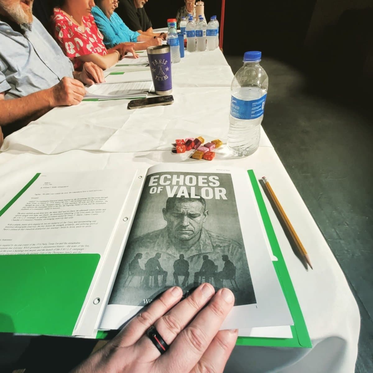 Cast members seated around a long table reading scripts at the first table read of Echoes of Valor, with the play cover visible in the foreground