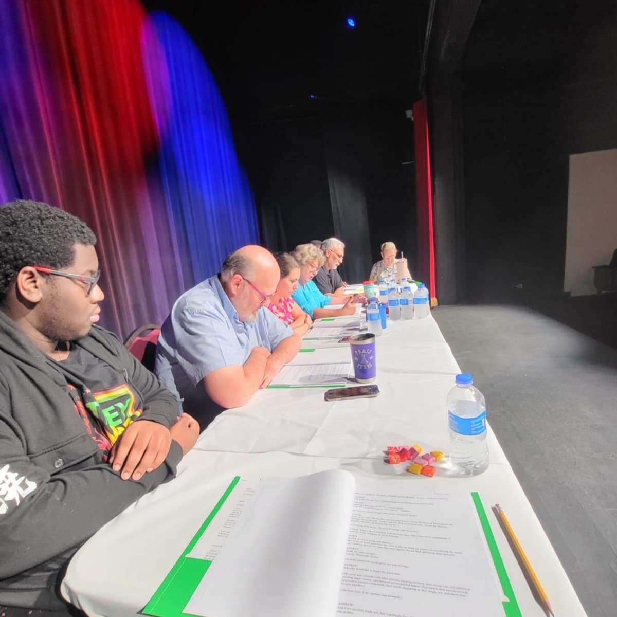 Wide shot of the full cast seated along a table reading scripts, with colorful stage curtains lit in red and blue behind them