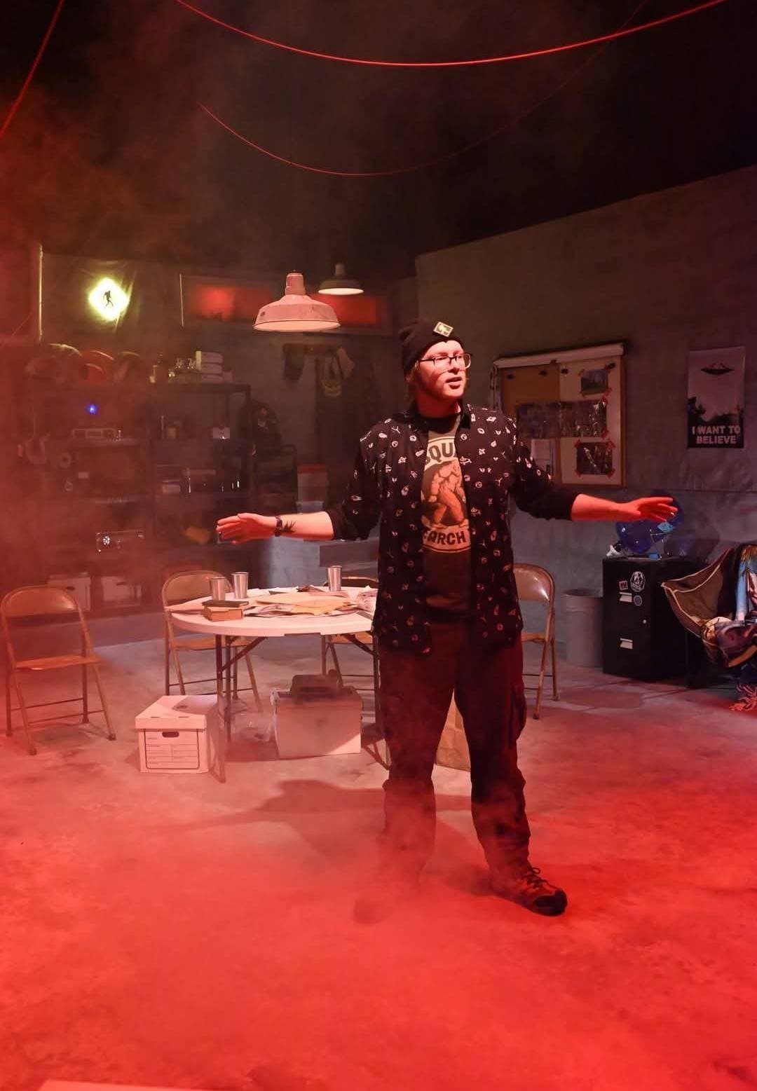 Actor playing Doug stands center stage with arms spread wide, surrounded by red-tinged fog, conspiracy board and I Want to Believe poster visible behind him