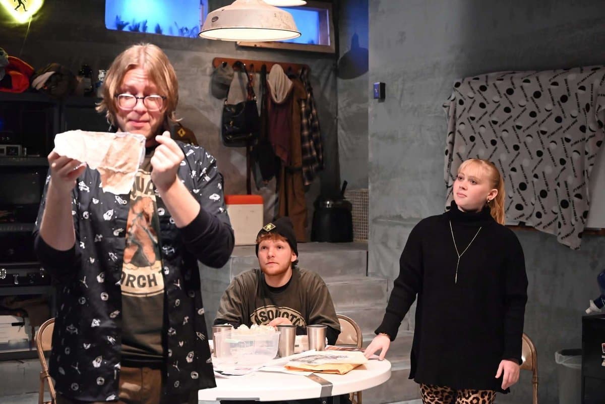 Doug holds up a mysterious piece of evidence while a male actor seated at the table and a female actor standing to the right look on skeptically