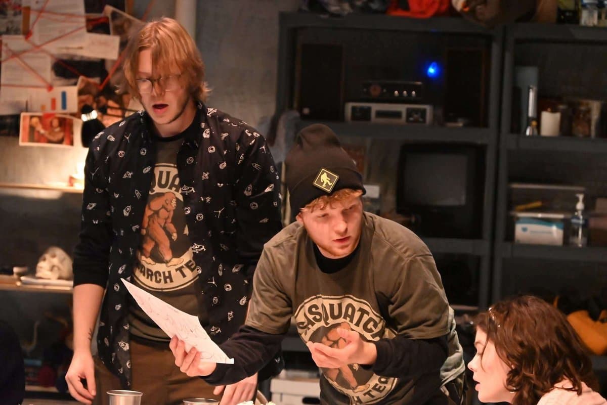 Doug and another male actor huddle over papers at the table while a female actor watches from the right, conspiracy board covered in red string visible behind them