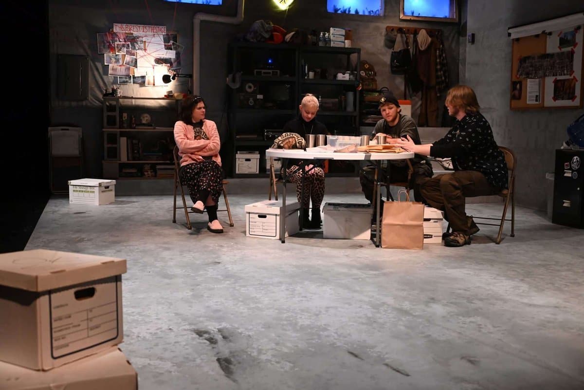Wide shot of all four actors seated around the table in the cluttered conspiracy-filled basement set, storage boxes scattered across the stage floor