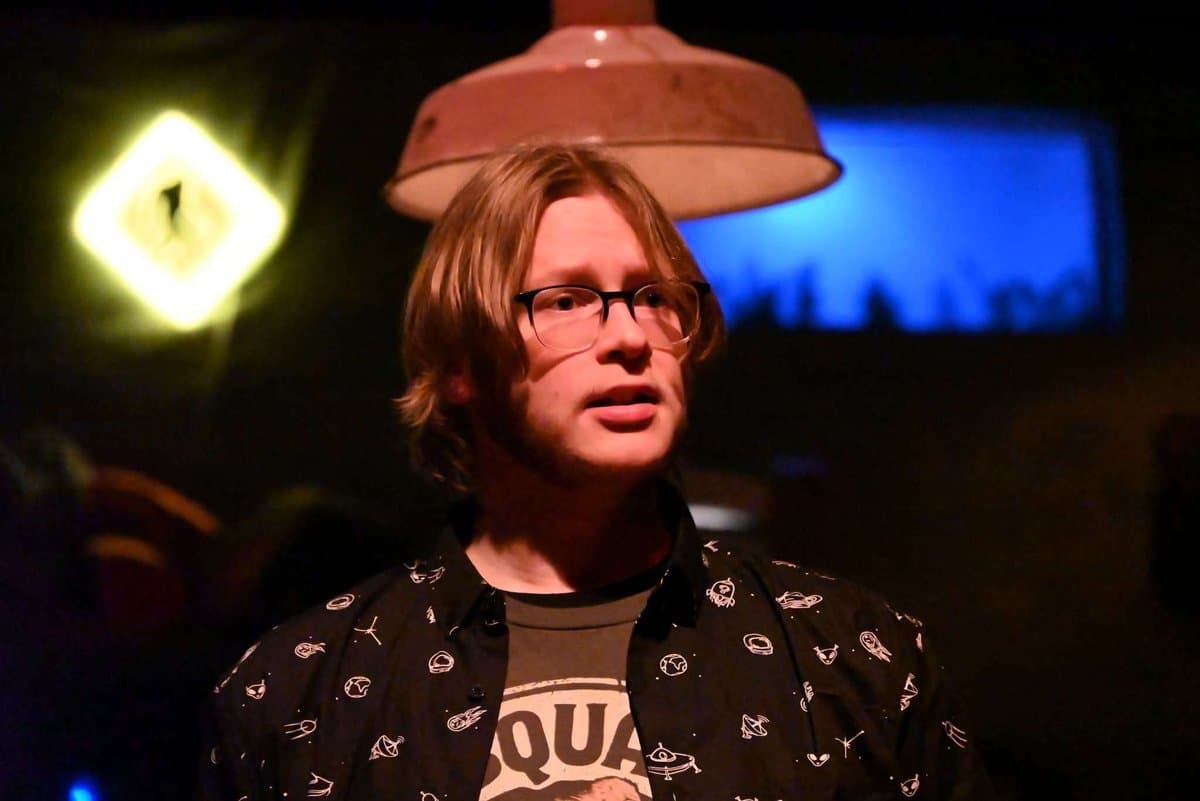 Close-up portrait of Doug under warm amber stage lighting, Sasquatch Research Team neon sign glowing yellow-green behind him